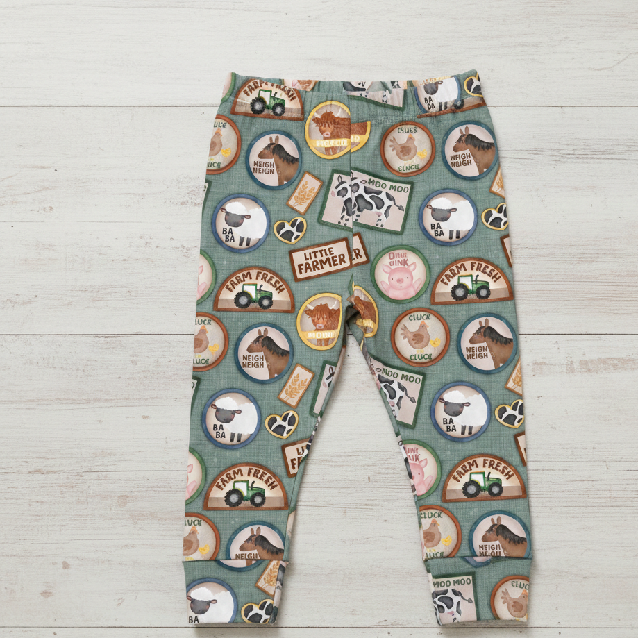 Children's light green leggings with farm-themed design including horses, tractors and sheep photographed on a white wooden surface